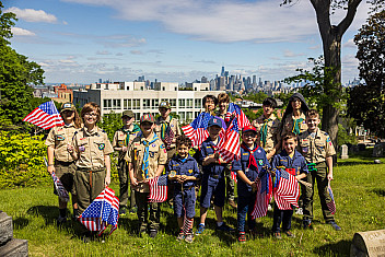 Scouts and Cub Scouts in uniform hold small American flags on a lawn in Green-Wood Cemetery, Brooklyn, with the Lower Manhattan skyline behind them, May 2025