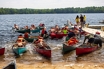 Scouts in life jackets push off in canoes from a sandy beach at Camp Yawgoog, Rockville, Rhode Island, during Troop 23 Brooklyn's summer camp week, July 2025