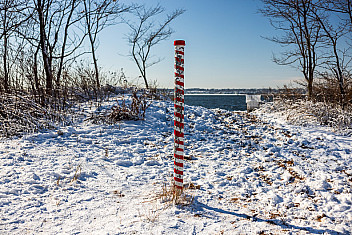 A red-and-white striped post on the snowy shore of Conference House Park, Staten Island, the marker NYC Parks calls the South Pole, at the southernmost point of New York State, January 2026