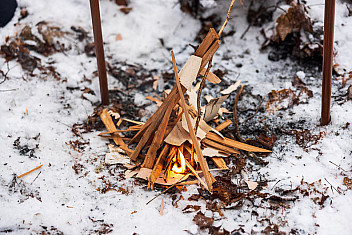 A small tepee of split kindling over wood shavings just catching fire in the snow, winter campout at Massawepie Scout Camps