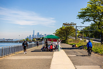 Walkers and a cyclist on the Shore Parkway Greenway in Bay Ridge, Brooklyn, with a volunteer tent beside the path, the 69th Street Pier and the lower Manhattan skyline across the harbor, May 16, 2026