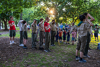 Scouts in uniform stand in a line and salute under the trees of Owl's Head Park in Bay Ridge, Brooklyn, with Cub Scouts and families behind them, on a June evening in 2024