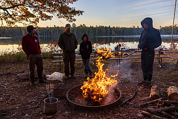 A campfire in a steel fire ring at dusk with people standing by and split firewood stacked nearby, Ten Mile River Scout Camps
