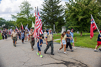 Scout leaders in uniform and a Cub Scout carrying a large American flag walk with families in the flag parade through Green-Wood Cemetery, Brooklyn, on the 250th anniversary of the Battle of Brooklyn, August 2026
