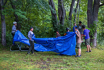 Troop 23 Brooklyn spreading a tarp between the trees at Lunksoos Camps after a day of riding in the rain, guyed with the twelve-foot paracord lengths the scouts had been practising knots on two days earlier, Thursday of Adventure Trek, August 13, 2026