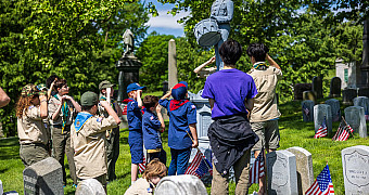Scouts and Cub Scouts salute at the statue of the drummer boy Clarence MacKenzie, among flagged soldiers' headstones in Green-Wood Cemetery, Brooklyn, May 2025