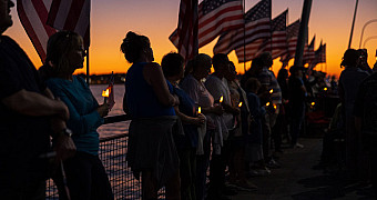 People hold candles along the railing of the American Veterans Memorial Pier at dusk under a row of American flags, Bay Ridge, September 11, 2025