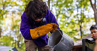 A Scout in a leather work glove pours water from a camp pot at the campsite, Camp Seton