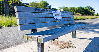 A weathered wooden bench with a notice taped to it on the Shore Parkway Greenway in Bay Ridge, before repainting in Jalen's Eagle Scout project, May 16, 2026
