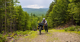 A rider pushing a loaded touring bike up the last of a steep gravel pitch, two more riders far below on the same ramp and the hills of Katahdin Woods and Waters behind them, Thursday of Troop 23 Brooklyn's Adventure Trek, August 13, 2026