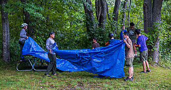Rigging a Tarp in the Rain โ Lunksoos Troop 23 Brooklyn spreading a tarp between the trees at Lunksoos Camps after a day of riding in the rain, guyed with the twelve-foot paracord lengths the scouts had been practising knots on two days earlier, Thursday of Adventure Trek, August 13, 2026