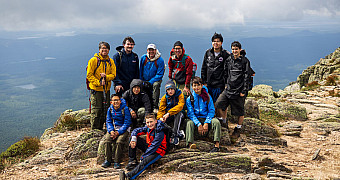 The whole party — scouts and adult leaders — on the summit ridge of Katahdin with the lakes and forest of Baxter State Park thousands of feet below, on the way down from Baxter Peak, Tuesday of Troop 23 Brooklyn's Adventure Trek, August 11, 2026