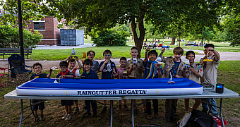 Boats Held High at the Raingutter Regatta Cub Scouts and Scouts hold up their sailboats behind the inflatable Raingutter Regatta course under the trees of Owl's Head Park, Bay Ridge, June 2024